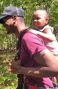 Customer Photo of a parent carrying a child with the Step Caddy child carrier - both smiling broadly. Step Caddy toddler carrier for outdoor adventures with kids, perfect for hiking and family outings.