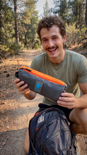 A smiling man outdoors on a forest path holds up a gray and orange zippered pouch with a blue logo patch. He is wearing an olive green t-shirt and a black backpack.