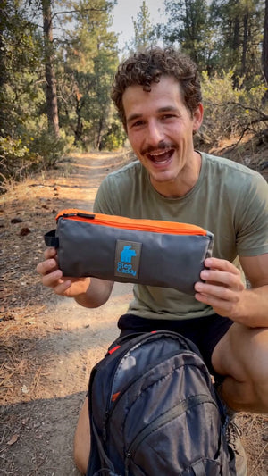 A man smiling in a forest while holding a gray and orange zippered pouch with a visible 'TERRA' logo patch. He is wearing an olive green t-shirt and a dark backpack.